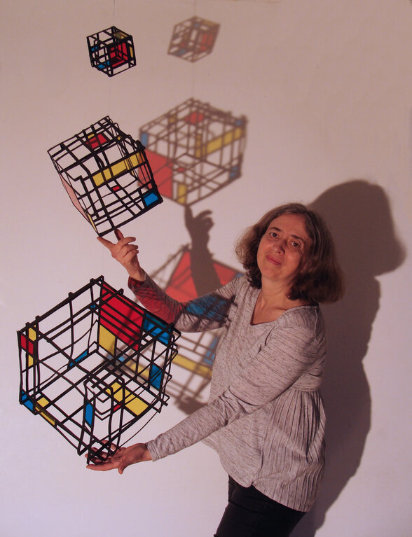 Une animatrice manipule des cubes filaires colorés dont les ombres géométriques projetées sur le mur créent un effet trompeur. - Agrandir l'image, fenêtre modale